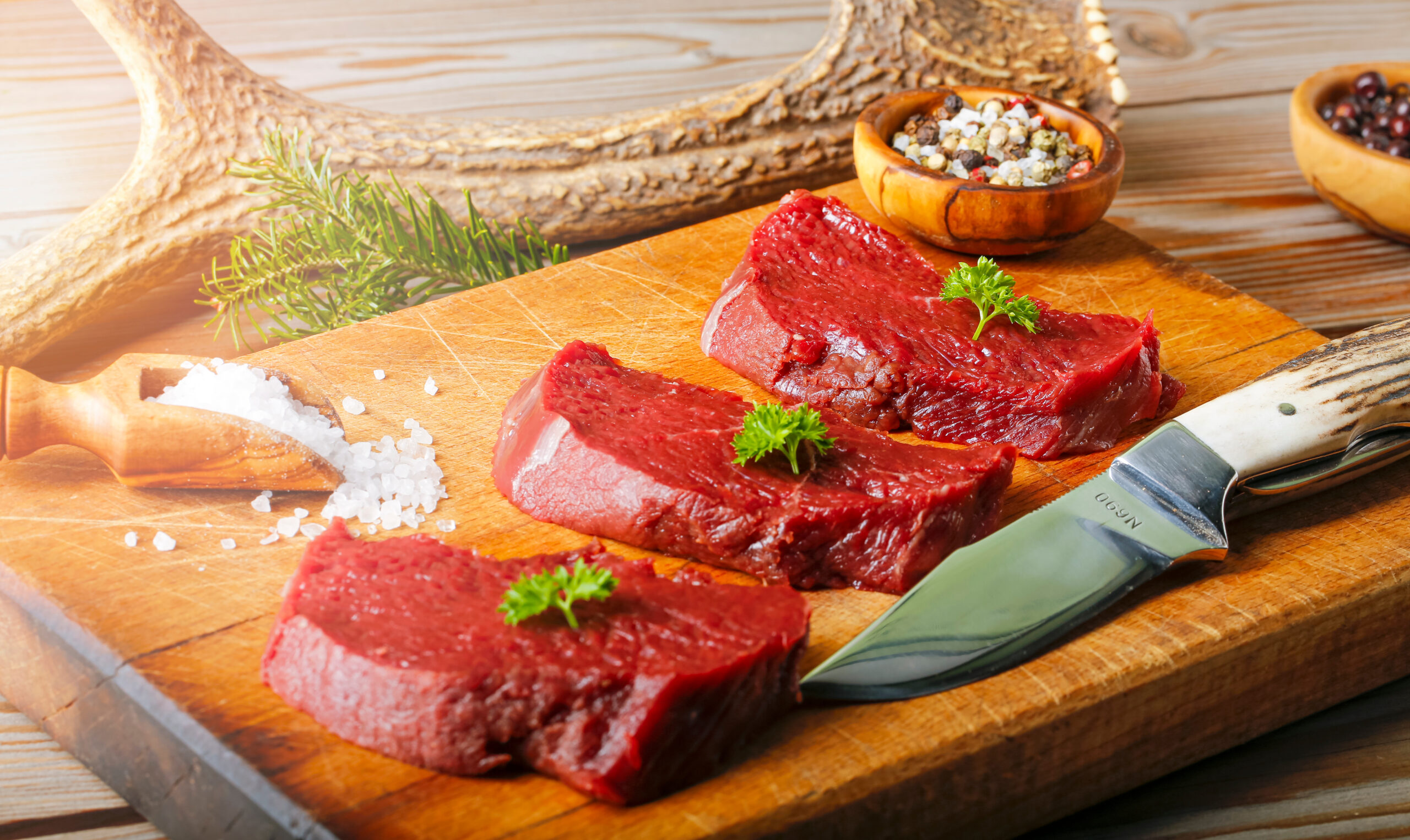 Raw meat from deer on the wooden chopping board with peppe. Hunting knife and deer antler as a decoration.